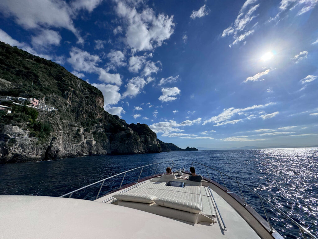 Two people on the bow of a boat cruising along Amalfi Coast cliffs under a sunny sky