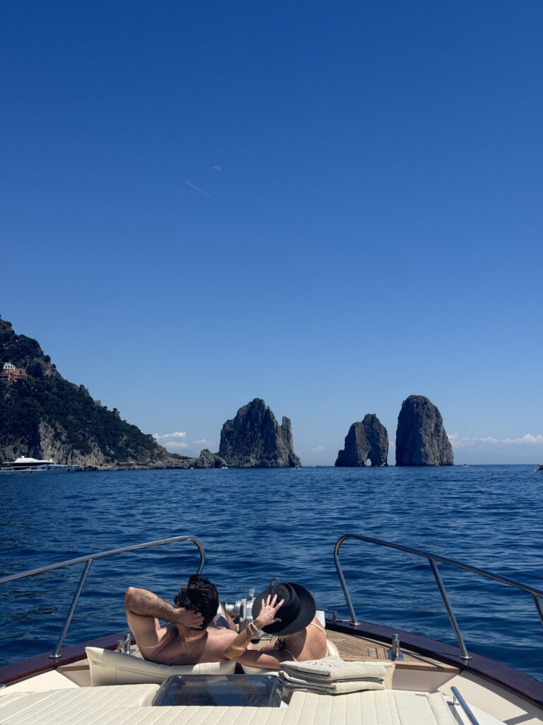View from the bow of a boat toward the Faraglioni rocks in Capri with two people relaxing