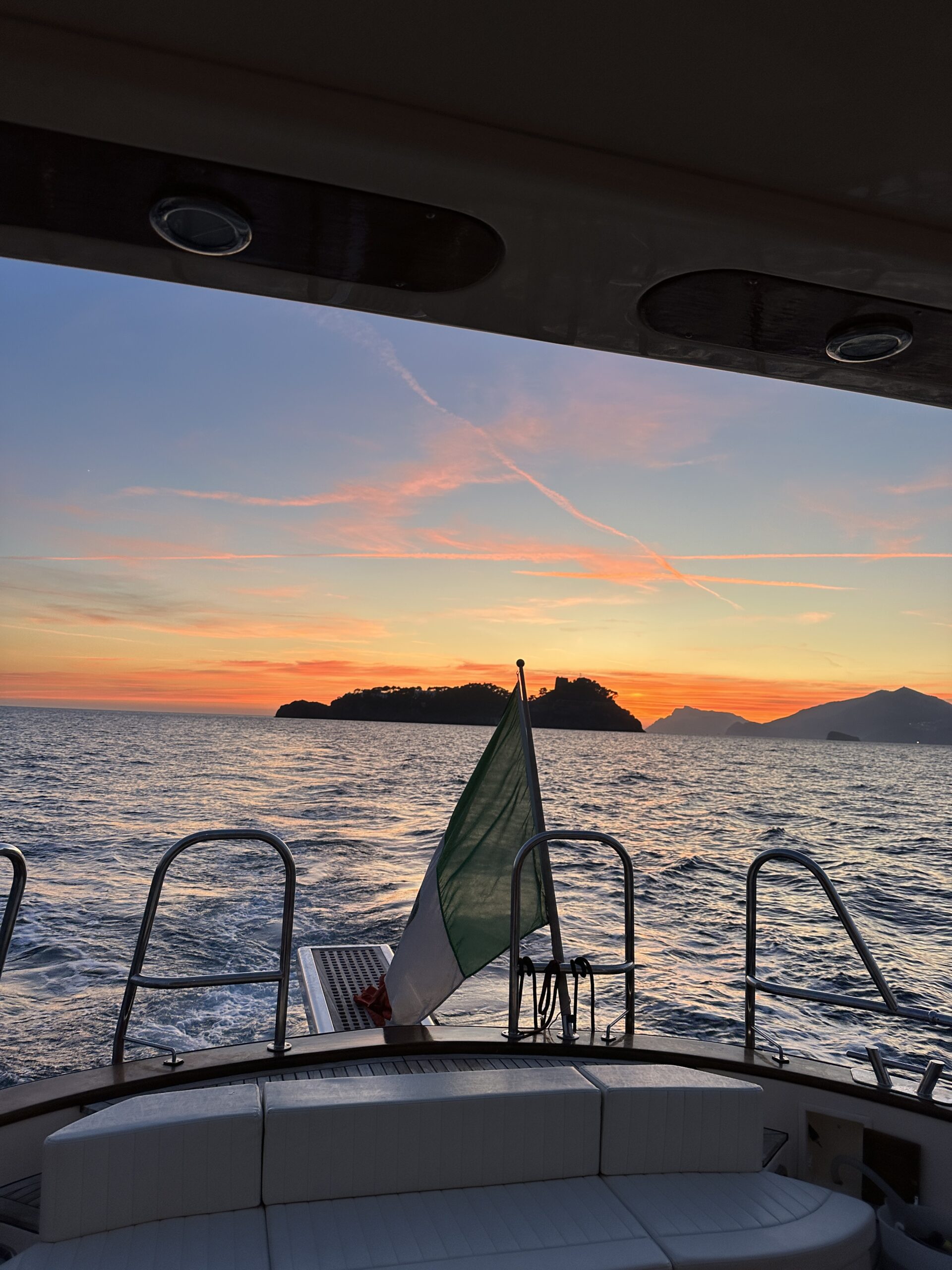 Sunset view from a boat with an Italian flag and island silhouette on the horizon